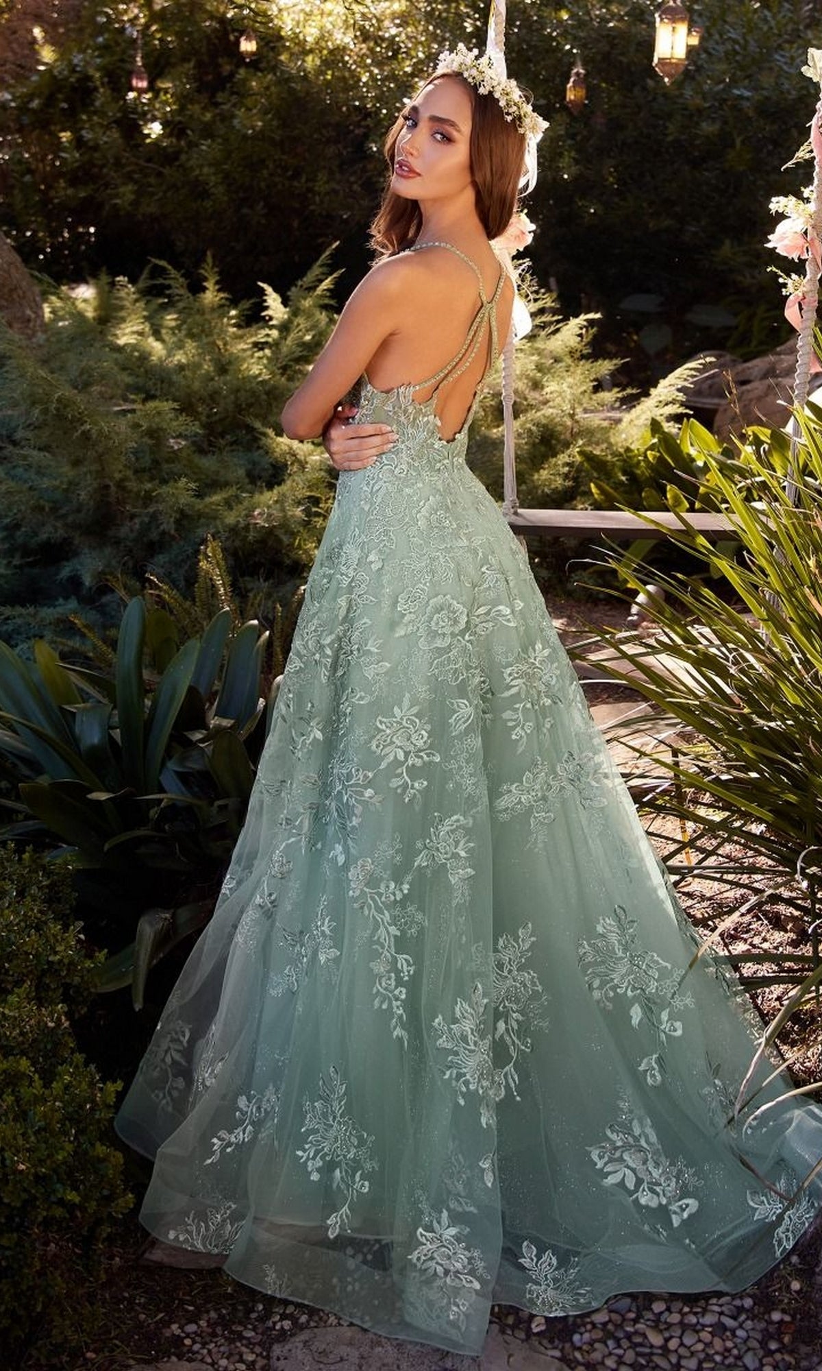 A woman stands outdoors in the Andrea & Leo A1248 Embroidered Prom Ball Gown—a light green, floor-length dress with thin straps and an open back—surrounded by greenery and decorative lights.